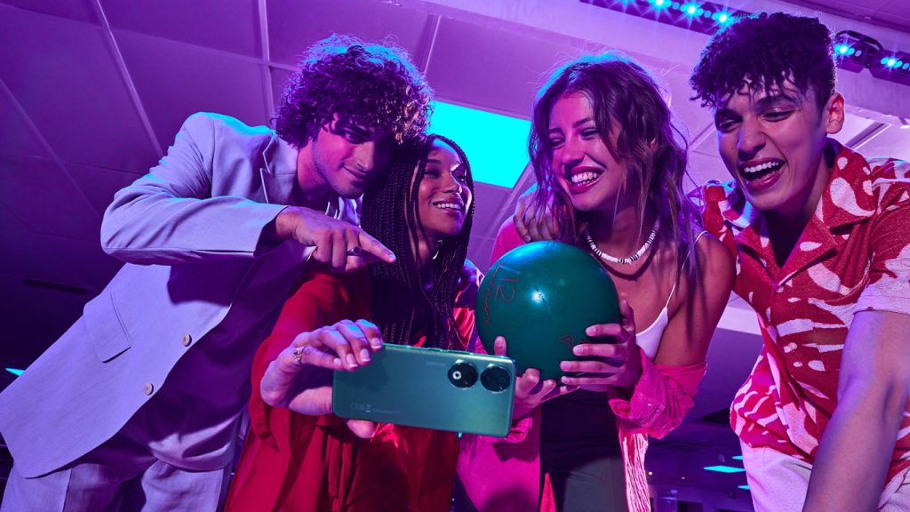 Skupina mladých ľudí hrá bowling a popri tom si robia selfie so smartfónom Honor 90
